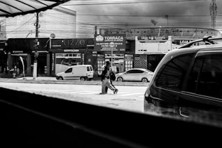 Person crosses street in front of shops and between cars.